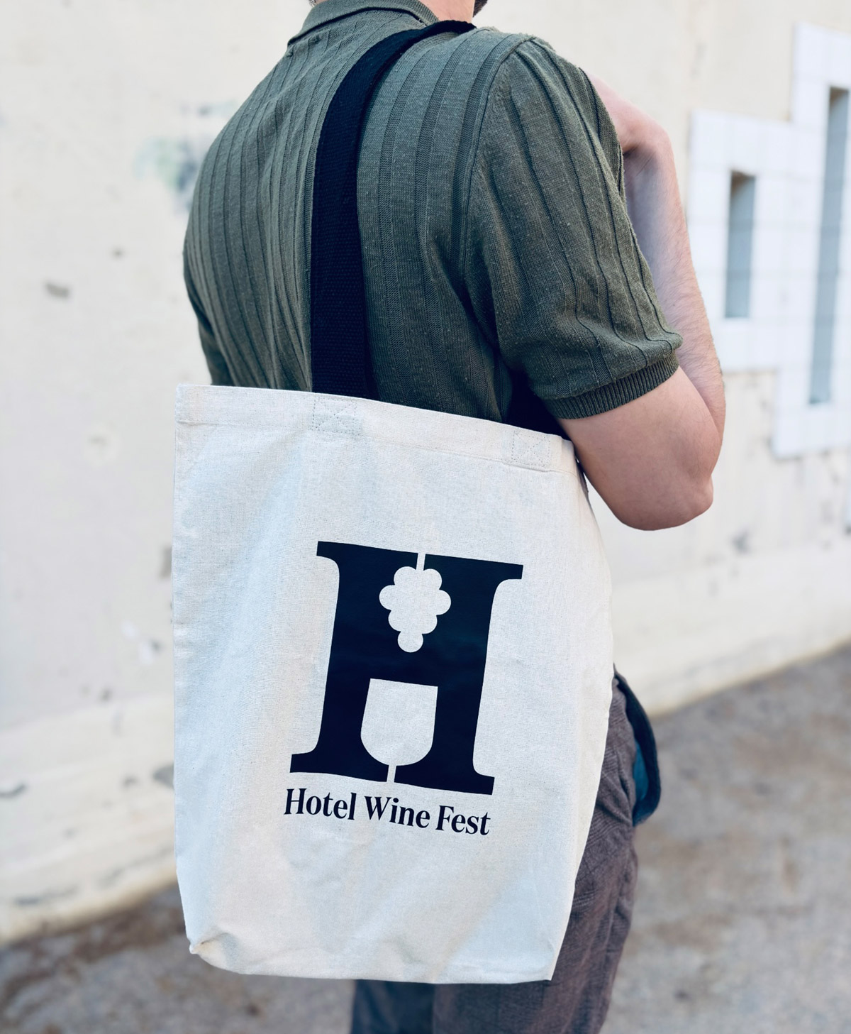 Tote bag de color blanco con el icono de la letra &ldquo;H&rdquo; de Hotel Wine Fest en color negro, aprovechando la contraforma de la propia letra para representar la silueta de un ramo de uvas y una copa de vino. En la parte inferior del icono se muestra el logotipo tipogr&aacute;fico de Hotel Wine Fest en una tipograf&iacute;a serif moderna.