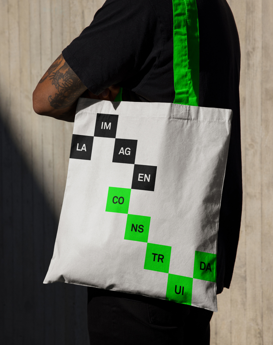 Tote bag en el hombro de un chico joven con camiseta corta negra, con la Campa&ntilde;a gr&aacute;fica para el ciclo de conferencias y debates, cuya primera edici&oacute;n se celebr&oacute; en La Virreina Centre de la Imatge en un sistema reticular que genera composiciones din&aacute;micas, donde palabras e im&aacute;genes se construyen y deconstruyen en cuadrados negros y verdes.