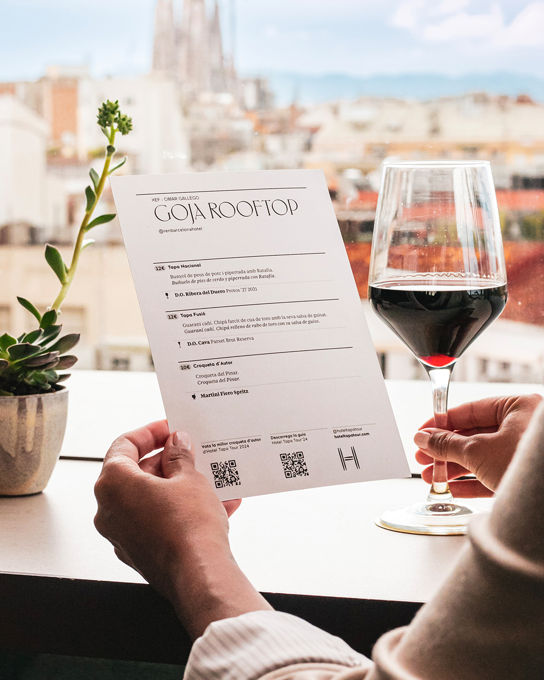 Cartas de restaurante con la identidad de Hotel Tapa Tour 2024 en una mesa con vistas a la ciudad de Barcelona. Una mujer sujetando con una mano la carta del restaurante, dise&ntilde;ada con una tipograf&iacute;a serif moderna y contempor&aacute;nea donde aparecen algunas de las tapa del restaurante Hotel Tapa Tour.