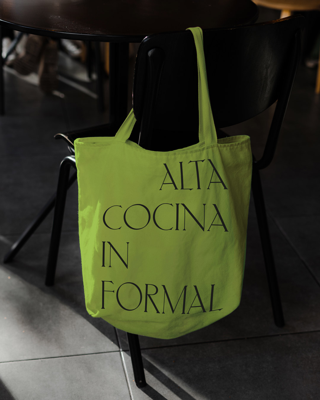 Tote bags verdes colgadas en una silla negra del interior de un bar. Contiene el eslogan de la nueva campa&ntilde;a e identidad de marca de Hotel Tapa Tour, en una composici&oacute;n tipogr&aacute;fica con justificaci&oacute;n alternada escrito con una tipograf&iacute;a serif moderna y contempor&aacute;nea que forma parte de la identidad de Hotel Tapa Tour de 2024.