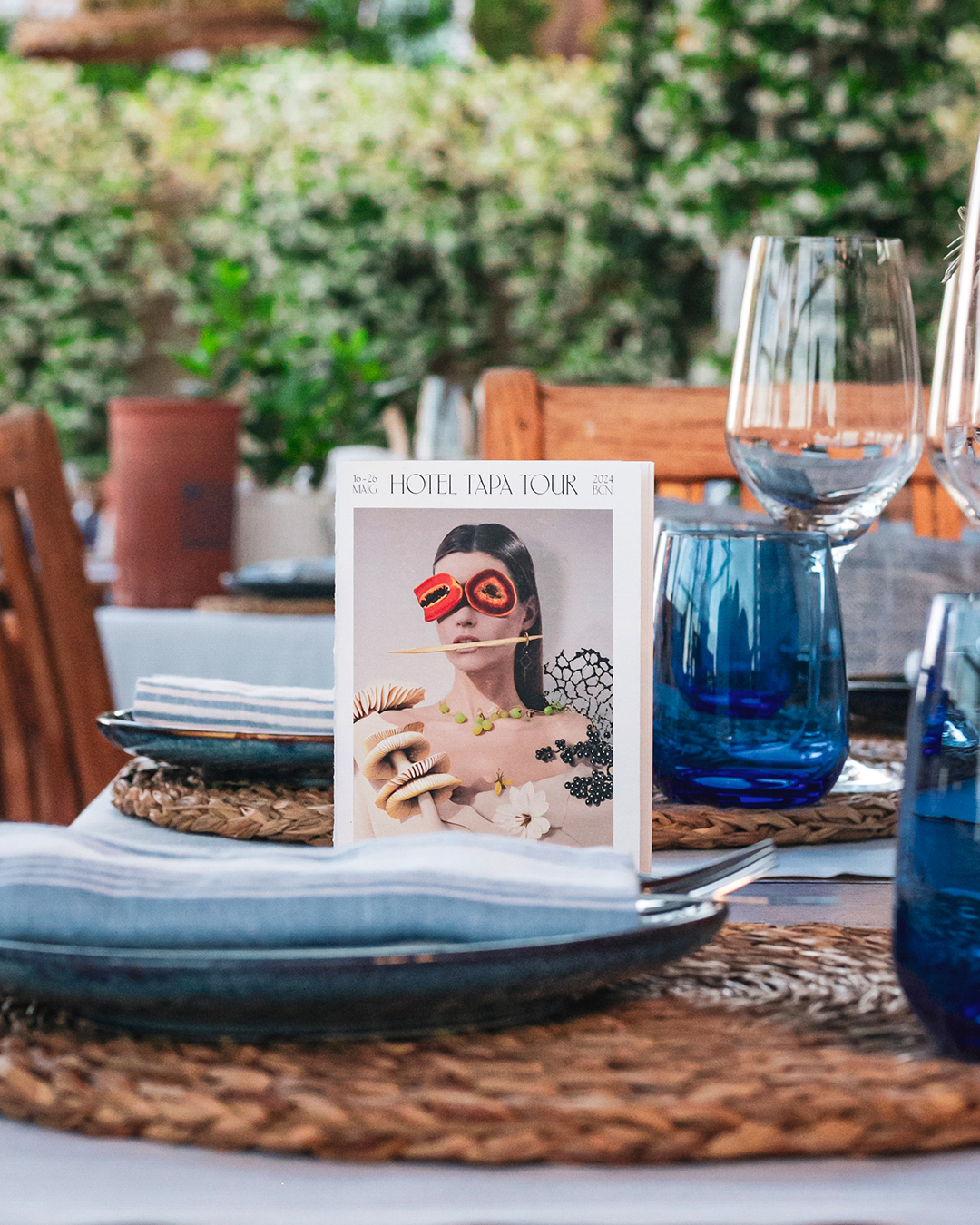 Cartas de restaurante con la identidad de Hotel Tapa Tour 2024 en una mesa al aire libre. Dise&ntilde;o vanguardista y transgresor con una tipograf&iacute;a serif moderna y contempor&aacute;nea reflejando la alta cocina informal de Hotel Tapa Tour. En el centro de la carta aparece la imagen de una chica con un vestido lleno de ingredientes que se integran en su vestuario creando un collage surrealista, consiguiendo una aut&eacute;ntica pieza de moda culinaria que forma parte de la misma campa&ntilde;a desarrollada por el estudio de dise&ntilde;o gr&aacute;fico Esiete.